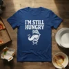 I'm Still Hungry distressed white text on a navy blue t-shirt with a graphic of a wide open mouth with