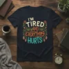 I'm Tired And Everything Hurts quote with floral accents on a navy t-shirt, humorous fatigue design