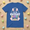 Blue t-shirt with the text "I'M THE CUTEST BUNNY" in colorful lettering, featuring bunny ears, feet, and decorated Easter eggs