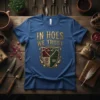 Gardening humor t-shirt with "In Hoes We Trust" text and a crest featuring garden tools, a rose, and a crown