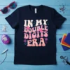 In My Double Digits Era text design with pink and orange wavy font and floral accents on a navy t-shirt