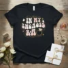 Black t-shirt with "IN MY ENGAGED R&A" in retro wavy font, surrounded by gold swirls, butterflies, and a diamond ring