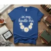 White text "in my kindle era" with daisy graphics on a blue t-shirt, featuring books and glasses icons