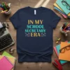 Navy t-shirt with "In My School Secretary Era" text in gold and teal glitter font, surrounded by small star and
