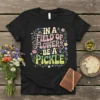 Black t-shirt with "In a Field of Flowers Be a Pickle" text in retro wavy font, surrounded by colorful flowers