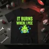 Black t-shirt with neon green dripping text "IT BURNS WHEN I PEE" and a green angry flame character with blue