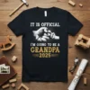 It is official I'm going to be a grandpa 2025 text with a fist bump graphic on a black t-shirt