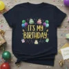Navy blue t-shirt with gold glitter text "IT'S MY BIRTHDAY" surrounded by balloons, cakes, and gift boxes