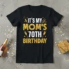 Black t-shirt with "IT'S MY MOM'S 70TH BIRTHDAY" in gold glitter and white text, featuring champagne glasses and music notes
