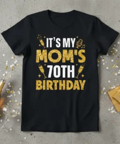 Black t-shirt with "IT'S MY MOM'S 70TH BIRTHDAY" in gold glitter and white text, featuring champagne glasses and music notes