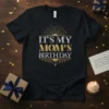 Black t-shirt with "IT'S MY MOM'S BIRTHDAY" text in gold and silver glitter font, art deco design
