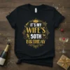 It's My Wife's 50th Birthday text with crown, champagne bottle, microphone, and glasses graphic on a black t-shirt