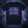 Black t-shirt with neon-style text reading "IT'S NOT EASY BEING MY FIANCEE'S ARM CANDY BUT HERE I AM NAILING IT"