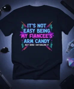 Black t-shirt with neon-style text reading "IT'S NOT EASY BEING MY FIANCEE'S ARM CANDY BUT HERE I AM NAILING IT"