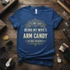 Navy blue t-shirt with gold text "IT'S NOT EASY BEING MY WIFE'S ARM CANDY BUT HERE I AM NAILING IT"