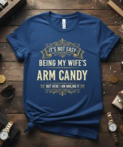 Navy blue t-shirt with gold text "IT'S NOT EASY BEING MY WIFE'S ARM CANDY BUT HERE I AM NAILING IT"