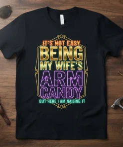 T-shirt with "It's Not Easy Being My Wife's Arm Candy But Here I Am Nailing It" quote in bold gold