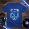 Blue t-shirt with white text "IT'S OKAY I'M WITH THE DJ" and neon blue DJ graphic with headphones and vinyl