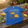 Blue t-shirt with "IT'S RAINING TACOS" text and a smiling taco holding an umbrella under a taco rain shower