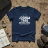 Navy blue t-shirt with "JOSHUA TREE NATIONAL PARK" and coordinates 33.874° N, 115.901° W in white and purple text, featuring
