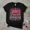 Black t-shirt with "JUICY PRINCESS SUPERSTAR" in glittery pink and silver text, adorned with crowns and swirls