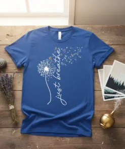White 'just breathe' text on a blue t-shirt with a dandelion graphic releasing seeds and butterflies
