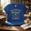 Blue t-shirt with gold text "Just Let Me Finish This Chapter" above an open book graphic with stars and constellations