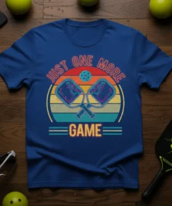 Blue t-shirt with "Just One More Game" text and graphic of crossed pickleball paddles and ball, featuring a retro sunset