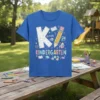 K Is For Kindergarten text with educational graphics like books, pencils, and globes on a blue t-shirt