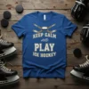 Keep Calm And Play Ice Hockey text with crossed hockey sticks and puck graphic on a royal blue t-shirt