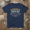 Navy blue t-shirt with "Keeper Of The Gender" text in gold and blue, featuring baby feet graphic