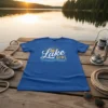 Blue t-shirt with "Lake Girl" text and sun graphic, set on a wooden dock by a lake at sunset