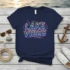 Retro graphic tee with "LAKE VIBES" text in bold leopard print and neon accents, featuring small pine trees and stars
