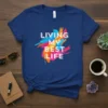White text "LIVING MY BEST LIFE" over colorful abstract paint splatters on a blue t-shirt