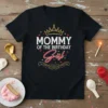 Mommy Of The Birthday Girl text with a crown graphic and decorative elements on a black t-shirt