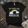 Black t-shirt with "MY FARTS ARE MAGICAL" text above a colorful rainbow and clouds, with glitter accents