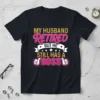 Black t-shirt with "MY HUSBAND RETIRED BUT HE STILL HAS A BOSS" text in pink and gold, featuring thumbs up