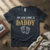 My New Name Is Daddy text with decorative font and baby feet graphic on a dark t-shirt