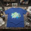 Nah graffiti spray paint design on a blue t-shirt, featuring white and green lettering with blue and green splatters