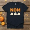 Black t-shirt with orange pixelated "NOM" text above three white dollops of whipped cream, part of a fall-themed design