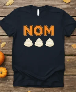 Black t-shirt with orange pixelated "NOM" text above three white dollops of whipped cream, part of a fall-themed design
