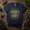 Navy blue t-shirt with "OLDER THAN DIRT" text in distressed gold font, surrounded by ornate green and brown circular design