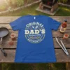 Original DAD'S Backyard BBQ AUTHENTIC text design with grill tools and flames on a blue t-shirt