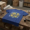 Blue t-shirt with distressed yellow text "PACKAHOLIC" and surrounding box graphics and arrows, on a wooden table with shipping supplies