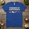 Blue t-shirt with white and gold text "POWERED BY KETONES" and graphics of steak, eggs, chicken leg, and bacon