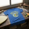 Blue t-shirt with yellow text "Pretty Pretty Pretty Good" above a cartoon sun character surfing a wave