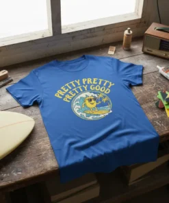 Blue t-shirt with yellow text "Pretty Pretty Pretty Good" above a cartoon sun character surfing a wave