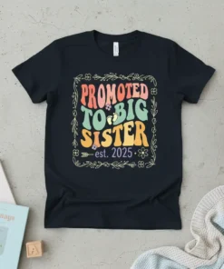 Promoted To Big Sister est. 2025 text with baby feet graphic on a black t-shirt, framed by floral elements