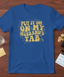 Blue t-shirt with gold retro lettering that reads "PUT IT ON. ON MY HUSBAND'S TAB", surrounded by champagne glasses and
