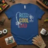 T-shirt with "REEL COOL DAD" text in blue, yellow, and red, featuring a fishing rod and a jumping bass graphic